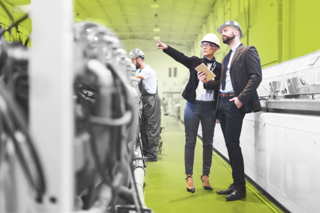 symbolic image a woman and a man are standing in a production hall she points to something further back he follows her finger pointing with his eyes both are wearing a helmet in the background there is a worker at a production station